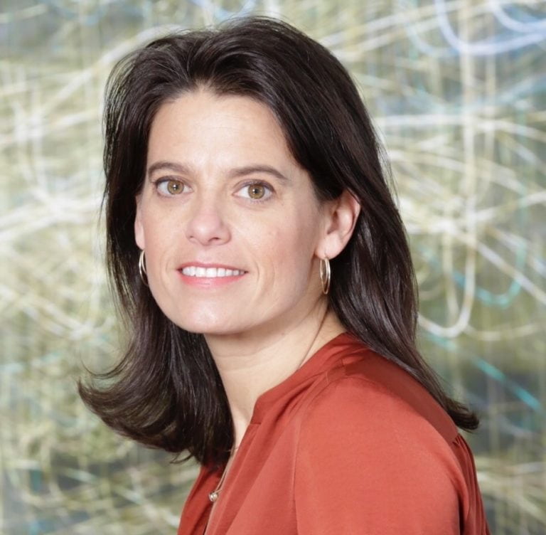 Woman with long dark hair, wearing a rust-colored top and hoop earrings, smiling slightly. The background is abstract with a mix of light lines and colors.