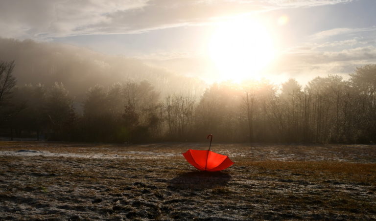 A bright red umbrella sits open on a frosty field at sunrise. Mist hovers over distant trees and hills, while the sun casts a warm glow across the landscape, creating a serene and atmospheric scene.