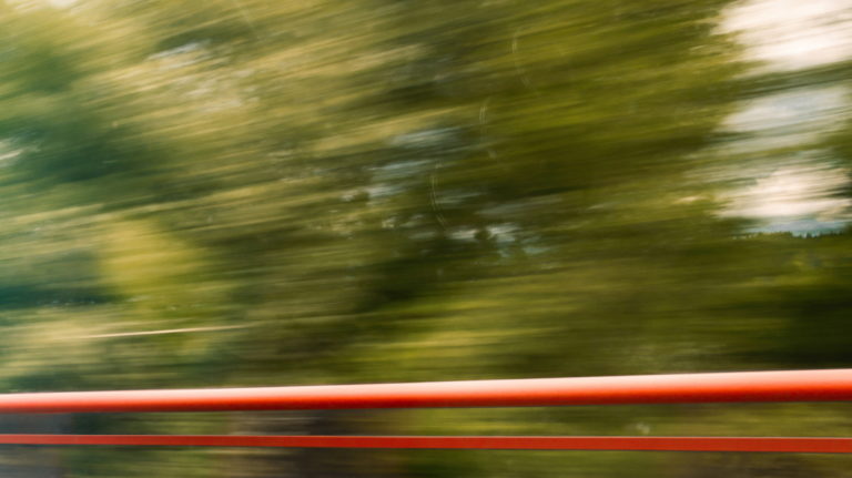 Blurred image of trees and greenery rushing past, with a red horizontal line in the foreground, suggesting movement. The scene conveys a sense of speed and motion, possibly taken from a moving vehicle.