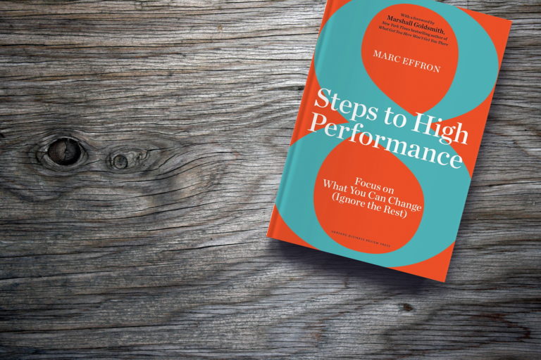 Book titled "Steps to High Performance" by Marc Effron rests on a wooden surface. The cover is orange with turquoise circles and text in white, featuring a subtitle: "Focus on What You Can Change (Ignore the Rest).