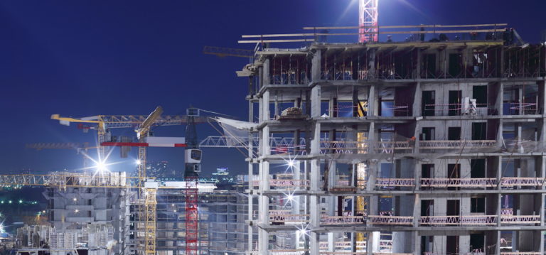 A nighttime construction site with partially built high-rise buildings and several illuminated cranes. The city lights are visible in the background, highlighting the urban setting.