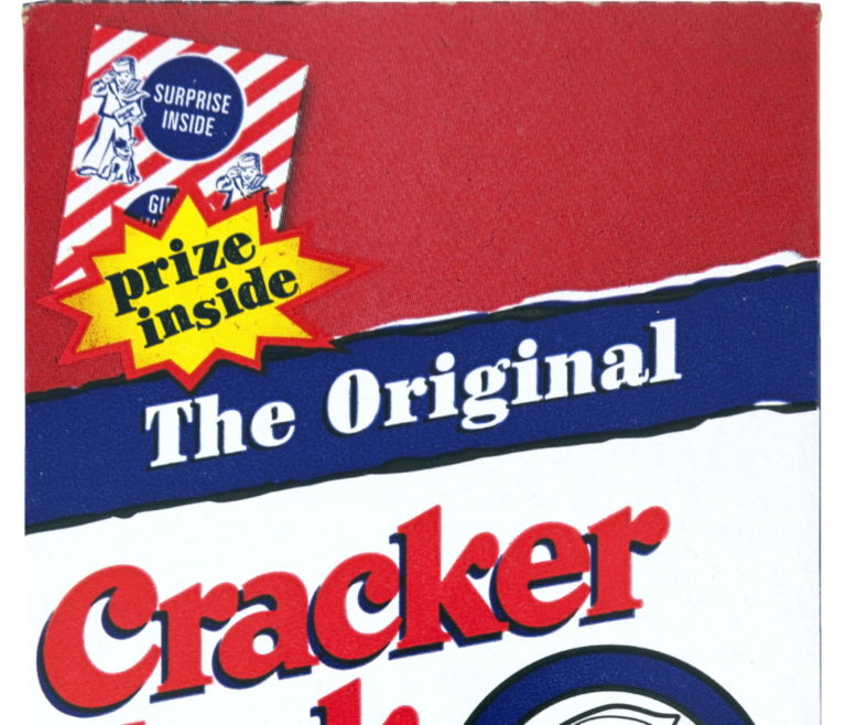 Close-up of a Cracker Jack box. The design includes the tagline "The Original Cracker Jack" with a yellow starburst stating "prize inside" and a striped corner with "Surprise Inside." The box features nautical-themed graphics.