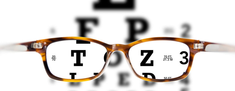 A pair of eyeglasses with a brown frame focus on an eye chart in the background. The chart has letters like E, F, P, T, and Z, which are clear through the lenses but blurry outside them.