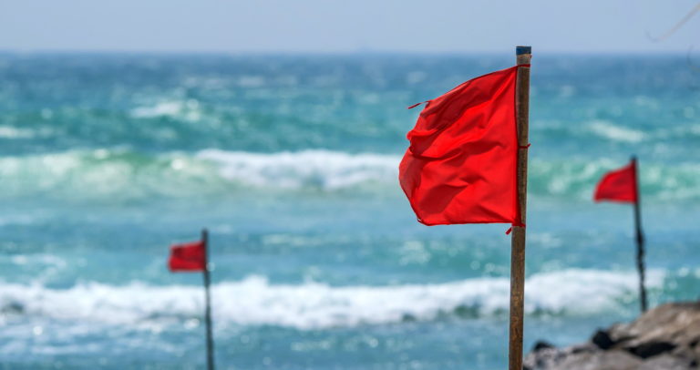 Red flags are mounted on poles along a beach, blowing in the wind against a backdrop of a turbulent blue sea and a clear sky, indicating caution due to dangerous conditions.