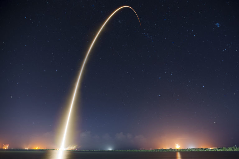 A long-exposure photograph of a rocket launch at night, showing a bright arc of light ascending into a starry sky. The scene is reflected on calm water, with the horizon faintly lit.