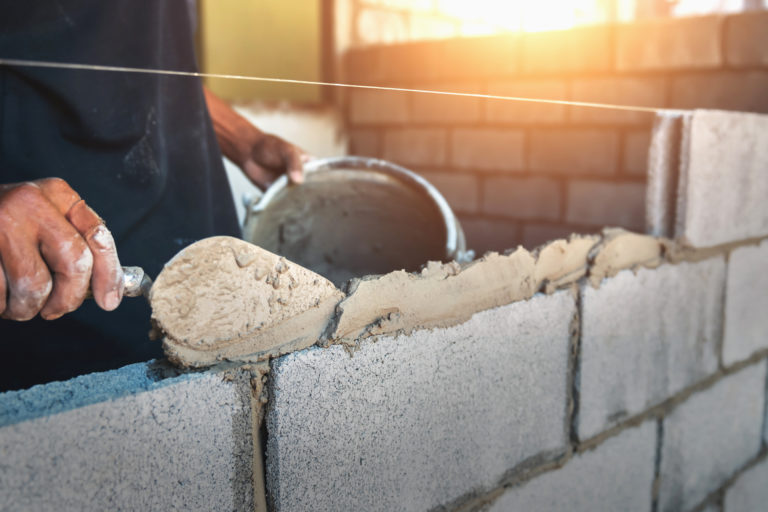 A person is applying mortar with a trowel to cement blocks while building a wall. The scene is well-lit, with sunlight streaming in from the background, highlighting the unfinished brickwork.