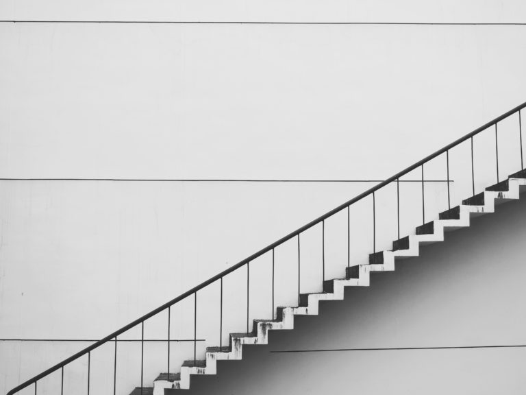 Black metal staircase with simple railing ascending diagonally on a minimalist white wall background, featuring sharp lines and geometric patterns.