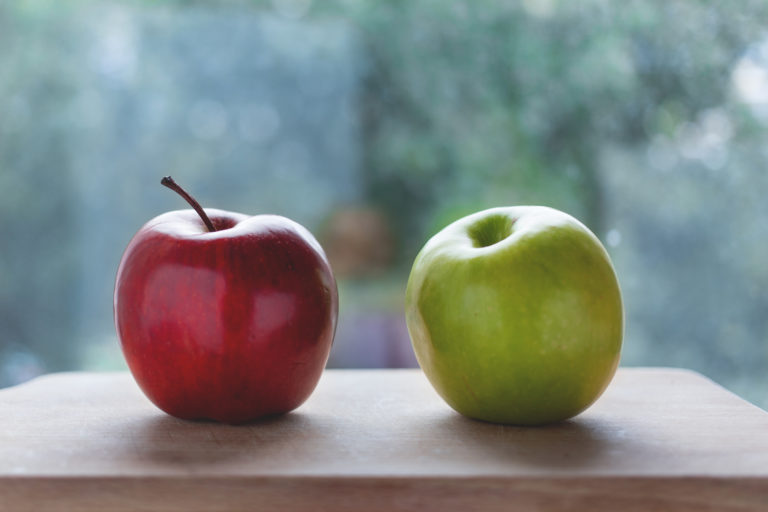 A red apple and a green apple sit side by side on a wooden surface, with a blurred outdoor background visible through a window. The apples are shiny and fresh, showcasing a contrast in color.