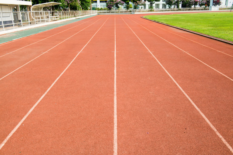 The image shows an empty red running track with white lanes curving around a sports field. There's a green grassy area to the right and bleachers on the left, surrounded by buildings in the background under a clear sky.