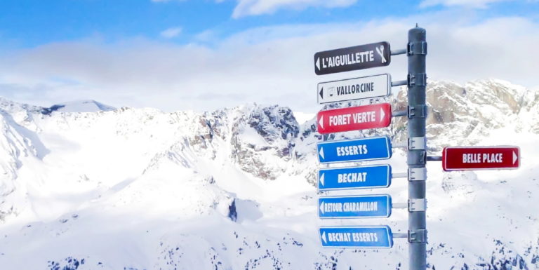 A snowy mountain landscape with a directional signpost in the foreground. The signpost points to various skiing routes with names like "L'Aiguillette," "Vallorcine," "Belle Place," and others. Snow-covered peaks and a blue sky are in the background.