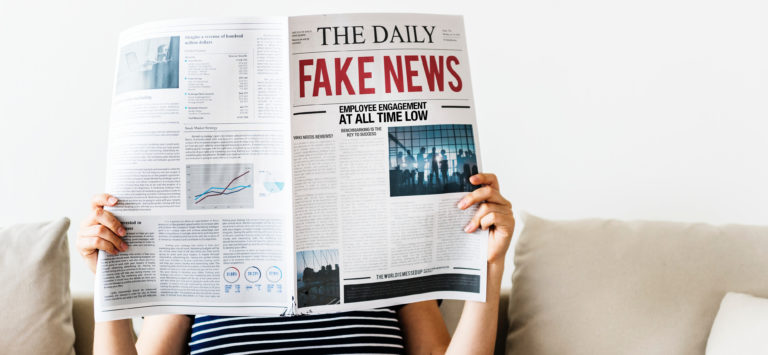 Person sitting on a couch reading a newspaper titled "THE DAILY," with "FAKE NEWS" in large red letters. The newspaper features various articles and charts. The person's face is obscured by the paper.