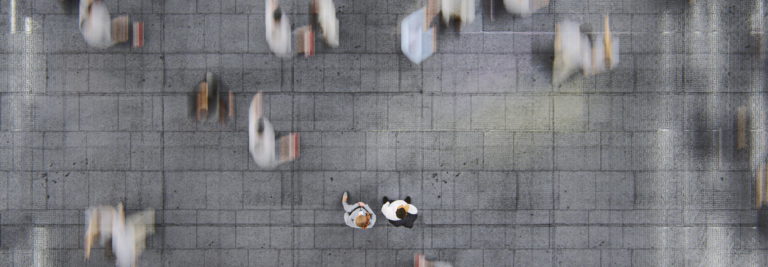 Overhead view of a busy sidewalk with blurred motion of people walking. Two individuals in the center stand still, one wearing white and the other black. The gray pavement is divided into square tiles.