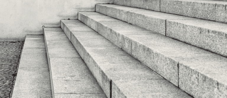 Close-up of wide, concrete steps leading up to an off-white wall. The surface is rough, and the steps have a slightly worn appearance. The scene is devoid of people, conveying a sense of stillness and simplicity.