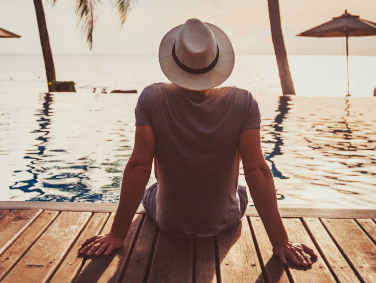 A person wearing a hat sits on a wooden deck by a pool, facing the ocean during sunset. Palm trees and umbrellas are visible in the background, creating a tropical and relaxing atmosphere.