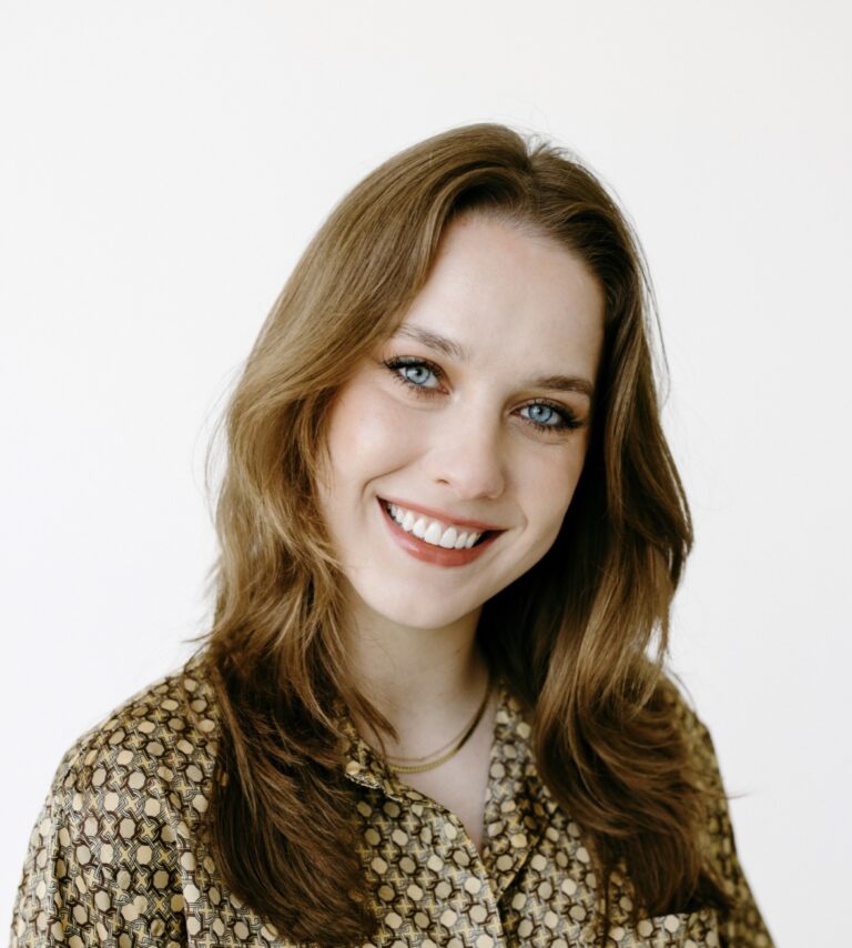 A smiling person with long, wavy brown hair is wearing a patterned blouse and a necklace. They are looking directly at the camera, set against a plain white background.