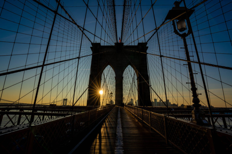 The sun sets behind the arches of the Brooklyn Bridge, casting long shadows on the pedestrian walkway. Cables fan out from the towers against the blue sky. Street lamps line the path, and cityscape silhouettes are visible in the distance.