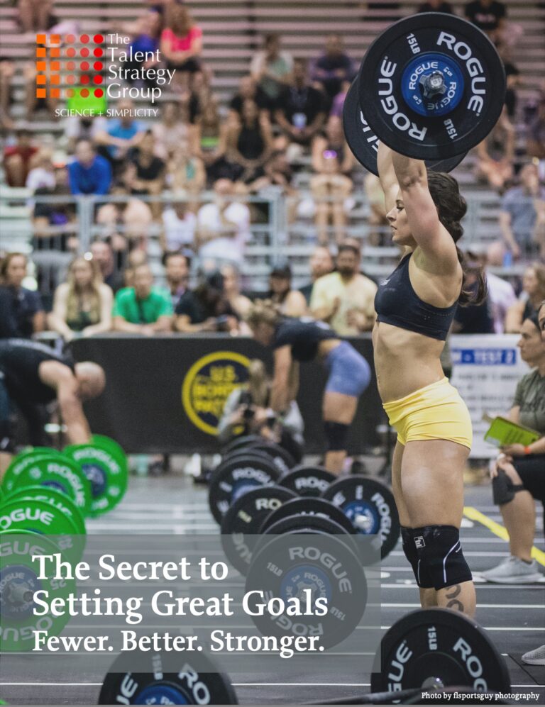 A woman in athletic gear lifts a barbell overhead in a gymnasium filled with spectators and competitors. The image has text reading, "The Secret to Setting Great Goals. Fewer. Better. Stronger." Gym equipment is visible in the background.